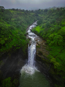 Aerial view of a tall waterfall cascading into a pool, surrounded by lush green hills and monsoon mist near Bhandardara, Maharashtra.
