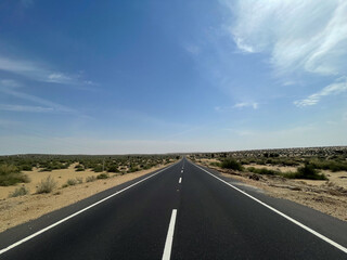 A long straight road cutting through the desert landscape near Longewala in Jaisalmer, India. Clear blue sky, open highway, and vast arid terrain create a dramatic scenic view.