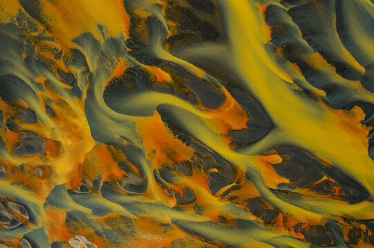 Aerial view of braided rivers carve through the landscape with vibrant hues of orange and yellow against the dark earth, South Coast, Iceland.