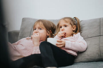 Two children, comfortably seated on a couch, are enjoying their snacks while watching a show, depicting warmth and familial togetherness in a cozy domestic setting.