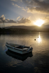 Fototapeta premium A moored small wooden fishing boat at dawn with sunlight breaking through the distant hills to cast light shallow on the calm water.