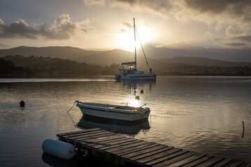 Fototapeta premium A wooden jetty with a small wooden boat, and a holiday sailing yacht in a sea channel with the morning sun rising over the hills in the distance.
