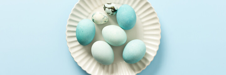 A collection of decorated blue Easter eggs sits on a white plate, showcasing various shades and patterns. The light background enhances the festive atmosphere of Easter, banner