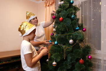 Mother and child decorate a Christmas tree in the room, wearing golden hats, cozy festive home atmosphere.
