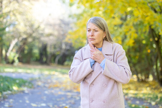 Senior woman standing outdoors in a park during autumn and experiencing severe toothache pain, touching her jaw and cheek with a distressed expression