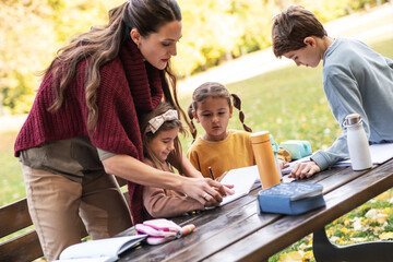 The teacher and children are now sitting together at a wooden table in the park, talking and observing nature around them. The moment is calm and focused, showing a gentle learning atmosphere outdoors