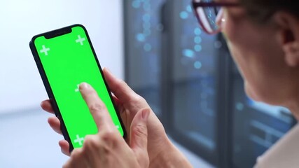 Closeup of a person holding a smartphone with a green screen tapping and swiping with a blurred background of a data center or server room ideal for technology and business concepts. - Powered by Adobe