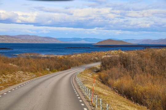 A winding road along Lake Tornetr&auml;sk in Lapland, northern Sweden. The deep blue water contrasts with the brown spring vegetation and the distant mountains under a partly cloudy sky.