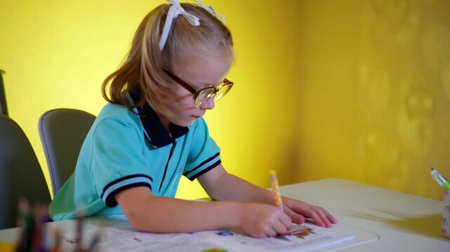 Focused child with light hair and chunky glasses concentrating on schoolwork at a white desk. Young student in a bright blue uniform intently writing in a workbook