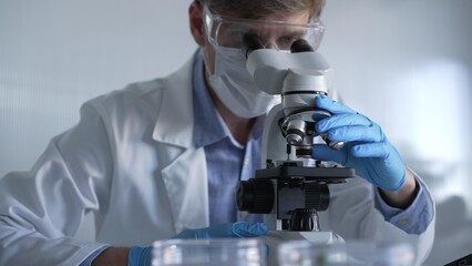 Male scientist researcher wearing blue gloves, protective glasses and medical mask, is using microscope in modern laboratory, portrait view. Low key lighting photo. Science and Medicine