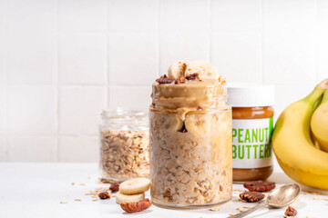 Peanut butter and banana overnight oatmeal, tasty sweet breakfast overnight oats in jar, with ingredients for cooking on kitchen table