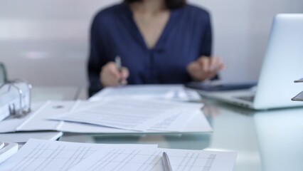 Magnifying glass, white calculator, pen and documents are lying on accountant's desk. Businesswoman wearing blue dress is working with documents on the background. Business and audit concept