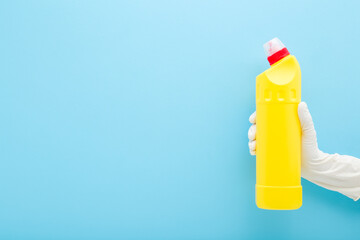 Woman hand in white rubber protective glove holding and showing yellow toilet cleaner bottle on blue wall background. Pastel color. Disinfection detergent. Closeup. Empty place for text. Front view.