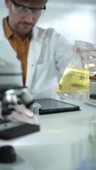 Man scientist wearing white protective gloves, and glasses is holding a yellow chemical solution...