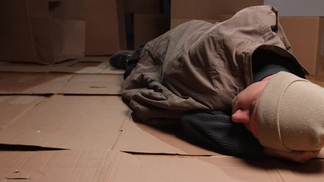 vagrant sleeps amid trash, unhoused individual finds shelter among broken materials and tattered clothing, homeless person takes refuge on shattered boxes surrounded by faded clothes and urban debris
