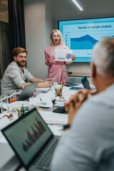 A diverse team gathers around a conference table as a woman in a pink suit presents charts and...
