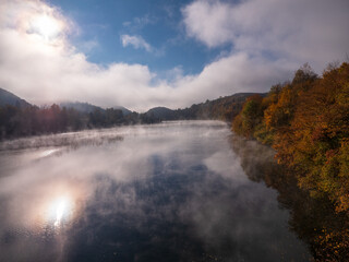 Fototapeta premium Morning mist over Ružín reservoir in Košice region, Slovakia. Autumn forest and sun reflection on calm water. Fog drifting above lake in peaceful mountain valley.