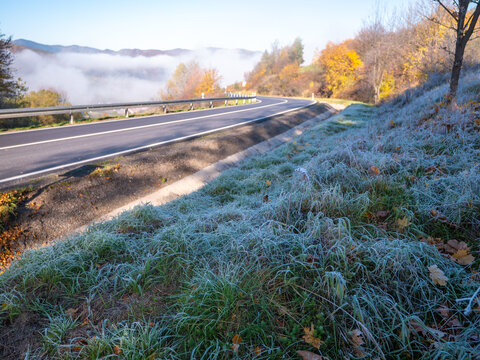 Frost-covered grass beside a mountain road on a clear autumn morning. First frost of the season, mist in the valley and colorful fall trees in the background.
