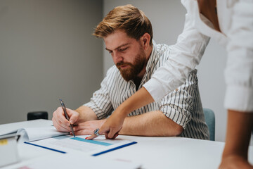 Focused teamwork as a colleague guides notes while a man writes on documents at a clean desk.
