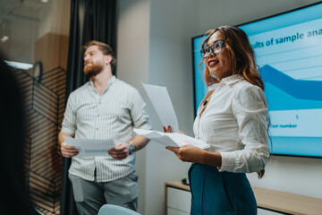 A confident woman in a white blouse and blue pants leads a presentation while a male colleague holds papers. A large screen displays a chart, highlighting teamwork and a productive office briefing.