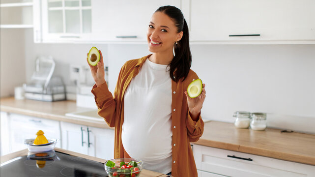 Happy pregnant lady showing two avocado halves while preparing healthy meal with lots of vegetables, standing at counter in kitchen, smiling at camera