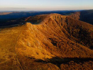 Aerial shot of Rothenbachkopf mountain ridge bathed in golden autumn morning sunlight, with soft shadows highlighting the rolling terrain of the Vosges mountains.