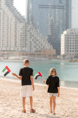 Fototapeta premium Abu Dhabi, United Arab Emirates. November 3, 2025: Two children hold UAE flags on the beach facing the water at Reem Island during National Day week