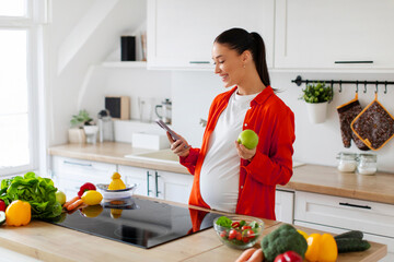 Smiling pregnant woman standing in modern kitchen holding apple in one hand and using cellphone...