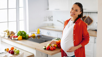 Portrait of young happy pregnant woman touching belly and smiling at camera, standing in kitchen. Healthy lifestyle and nutrition during pregnancy concept