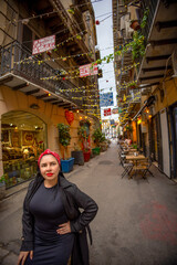 A confident woman poses in a charming Italian street decorated with lights and colorful signs. Cozy...