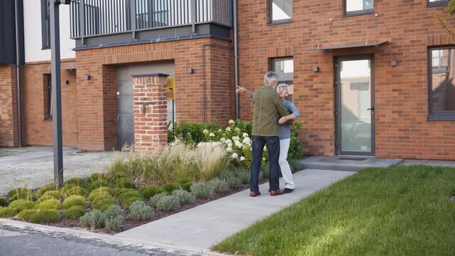 Gray haired man walking arm around mature woman along garden path toward brick house. Couple hugging close while moving together. Family enjoying moment arriving at modern home entrance.