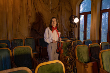 Woman in white blouse and checkered pants, holding violin, stands in old theater with green seats. Dramatic lighting from spotlight, autumn leaves scattered around. Red scarf adds vibrant touch