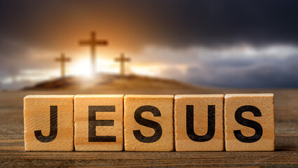 Row of wooden cubes with text of Jesus on the table with a Christian cross on the hill with dark dramatic sky background. Concept of Christianity, religious, faith, Jesus, or belief