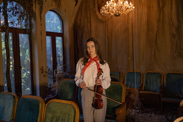 Elegant woman in white blouse and checkered pants holds violin in abandoned theater. Chandelier glows above, green velvet seats surround her. Autumn leaves and draped curtains add nostalgic touch