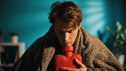 Young man with a fever suffering from seasonal flu, wrapped in a blanket and holding a hot water bottle to his chest to relieve chest pain and coughing while sitting at home at night