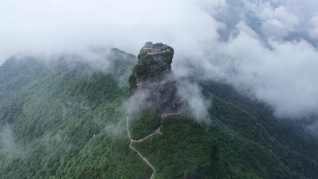 Aerial view of a majestic mountain peak covered in mist and lush green trees, with a winding pathway leading to the top, Fanjingshan, Guizhou, China.