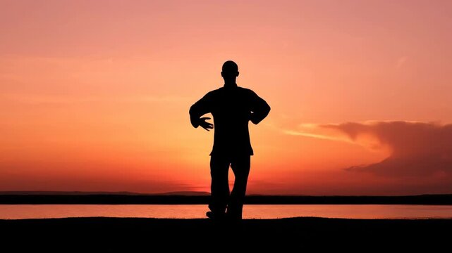 Silhouette of a person practicing tai chi at sunset. Vertical video of a stylized figure in a meditative qigong pose by the water. Wellness and balance concept