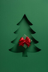A green holiday card features a layered conifer silhouette with a red ribbon gift at its center,...