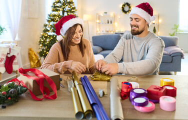 Christmas happy couple wrapping gift together near festive tree. In cozy living room, man and woman...