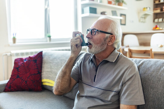 Senior man using asthma inhaler for relief an attack at home. Mature man using medical inhaler to prevent and treat wheezing and shortness of breath caused by allergy.