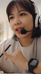 Close up of Young Asian woman working as a call center agent from home, speaking through a headset microphone while helping customers online.
