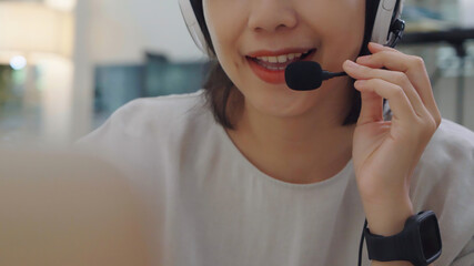 Young Asian female agent chatting through a headset microphone at home, offering kind customer assistance and staying relaxed during remote work.