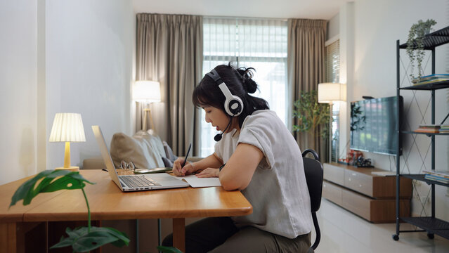 Young Asian woman wearing a headset taking notes during an online class at home, focused and learning with laptop.