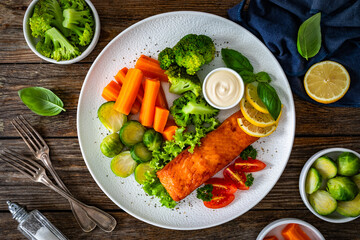 Fried salmon steak with boiled broccoli, brussels sprouts and carrots and mayonnaise on wooden table. Top view