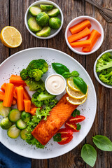 Fried salmon steak with boiled broccoli, brussels sprouts and carrots and mayonnaise on wooden table. Top view