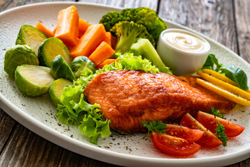 Fried salmon steak with boiled broccoli, brussels sprouts and carrots and mayonnaise on wooden table	