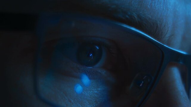 Close up of focused serious man eye wearing glasses illuminated by blue light from a computer or smartphone screen, working late at night or scrolling through a social media news feed.