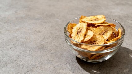 Crispy fried plantain chips fill a clear glass bowl resting on a textured gray surface.