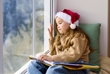 Christmas child in Santa hat reading by window. On a cozy windowsill the child pauses with an open...