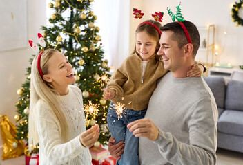 Christmas family with child holding sparklers indoors. Warm lights glow on the tree as they laugh...